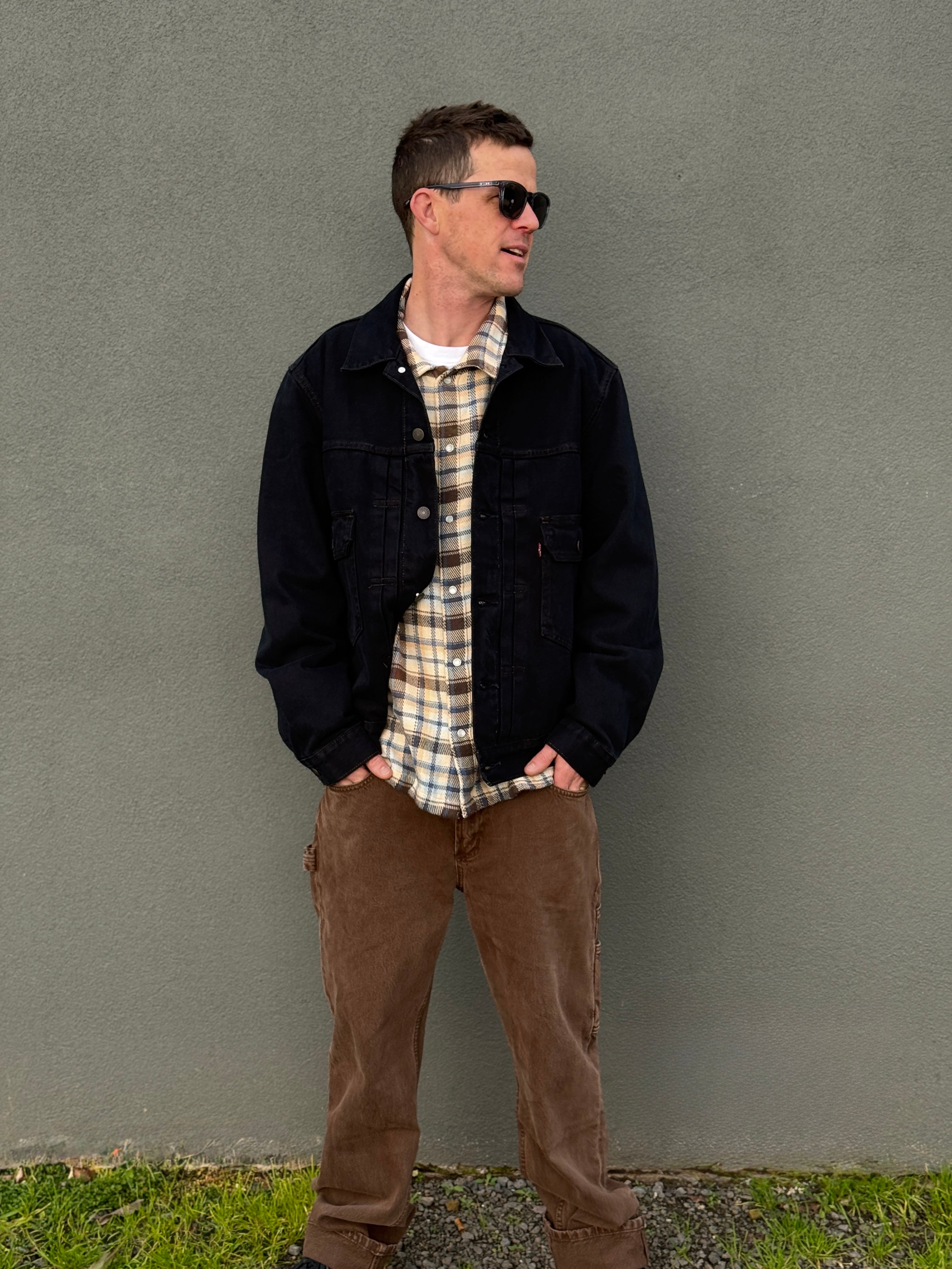 A man wearing a dark denim jacket over a beige and white plaid flannel shirt, paired with brown pants, standing against a textured gray wall.