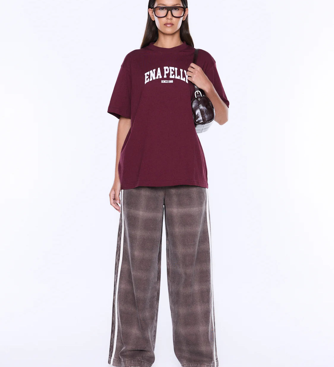 A model wearing an oversized maroon t-shirt with 'ENA PELLIS' printed in white on the chest, paired with wide-leg brown plaid pants featuring white side stripes, set against a plain white studio background.