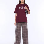 A model wearing an oversized maroon t-shirt with 'ENA PELLIS' printed in white on the chest, paired with wide-leg brown plaid pants featuring white side stripes, set against a plain white studio background.