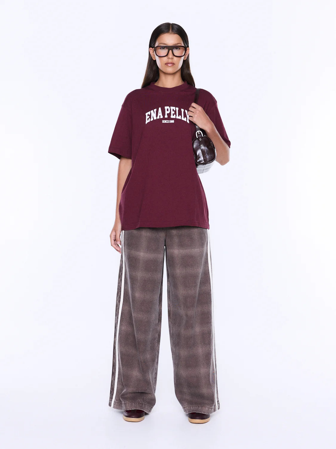 A model wearing an oversized maroon t-shirt with 'ENA PELLIS' printed in white on the chest, paired with wide-leg brown plaid pants featuring white side stripes, set against a plain white studio background.