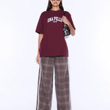 A model wearing an oversized maroon t-shirt with 'ENA PELLIS' printed in white on the chest, paired with wide-leg brown plaid pants featuring white side stripes, set against a plain white studio background.
