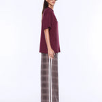 A woman modeling an oversized short-sleeve t-shirt in deep burgundy paired with wide-leg plaid trousers in brown and gray with a white stripe, set against a plain white studio background.