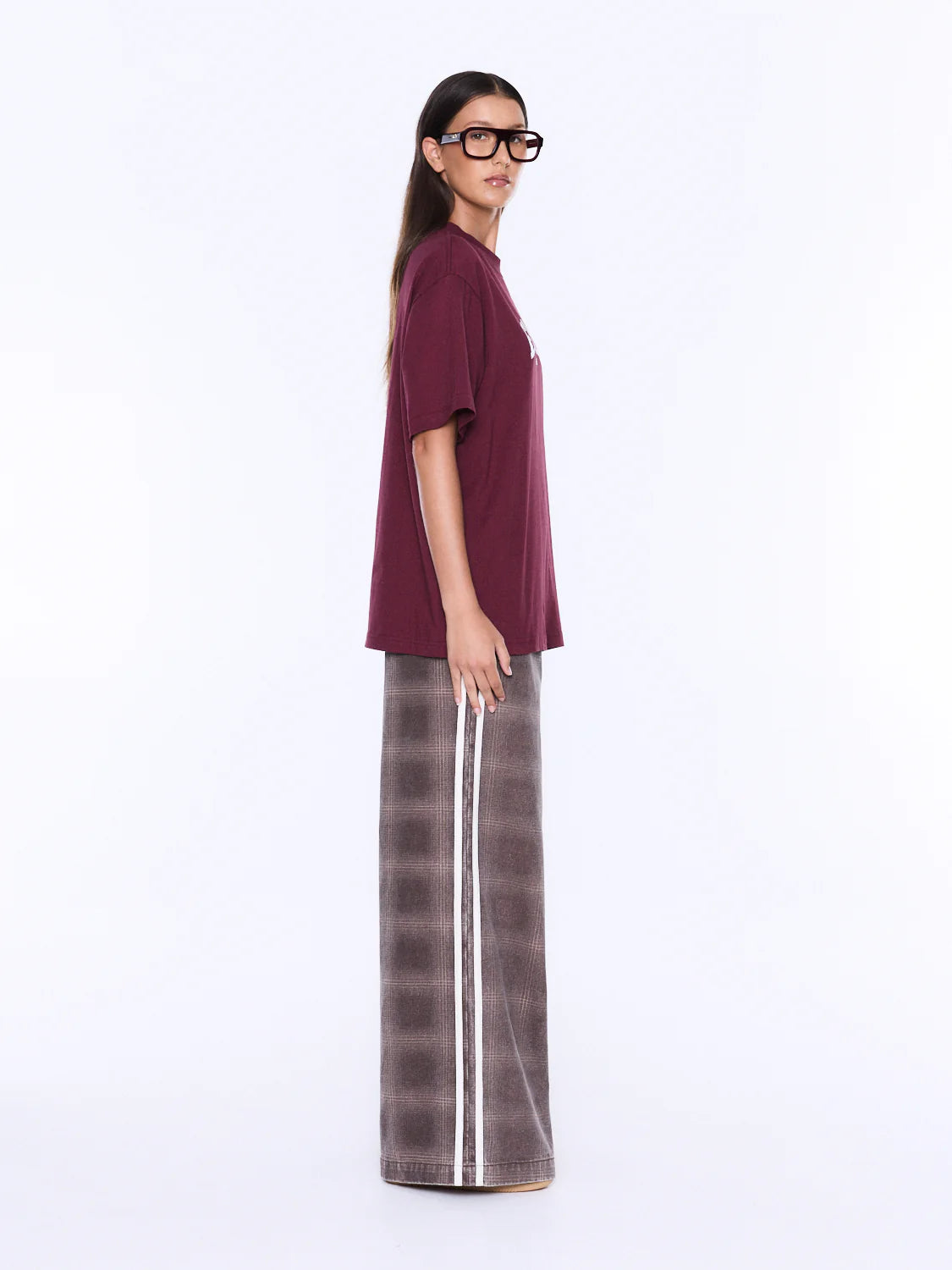 A woman modeling an oversized short-sleeve t-shirt in deep burgundy paired with wide-leg plaid trousers in brown and gray with a white stripe, set against a plain white studio background.