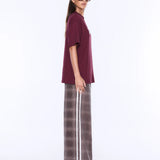 A woman modeling an oversized short-sleeve t-shirt in deep burgundy paired with wide-leg plaid trousers in brown and gray with a white stripe, set against a plain white studio background.
