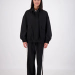 A woman stands against a light gray studio background wearing a black oversized zip-front jacket with front flap pockets and matching black wide-leg pants with white side stripes.