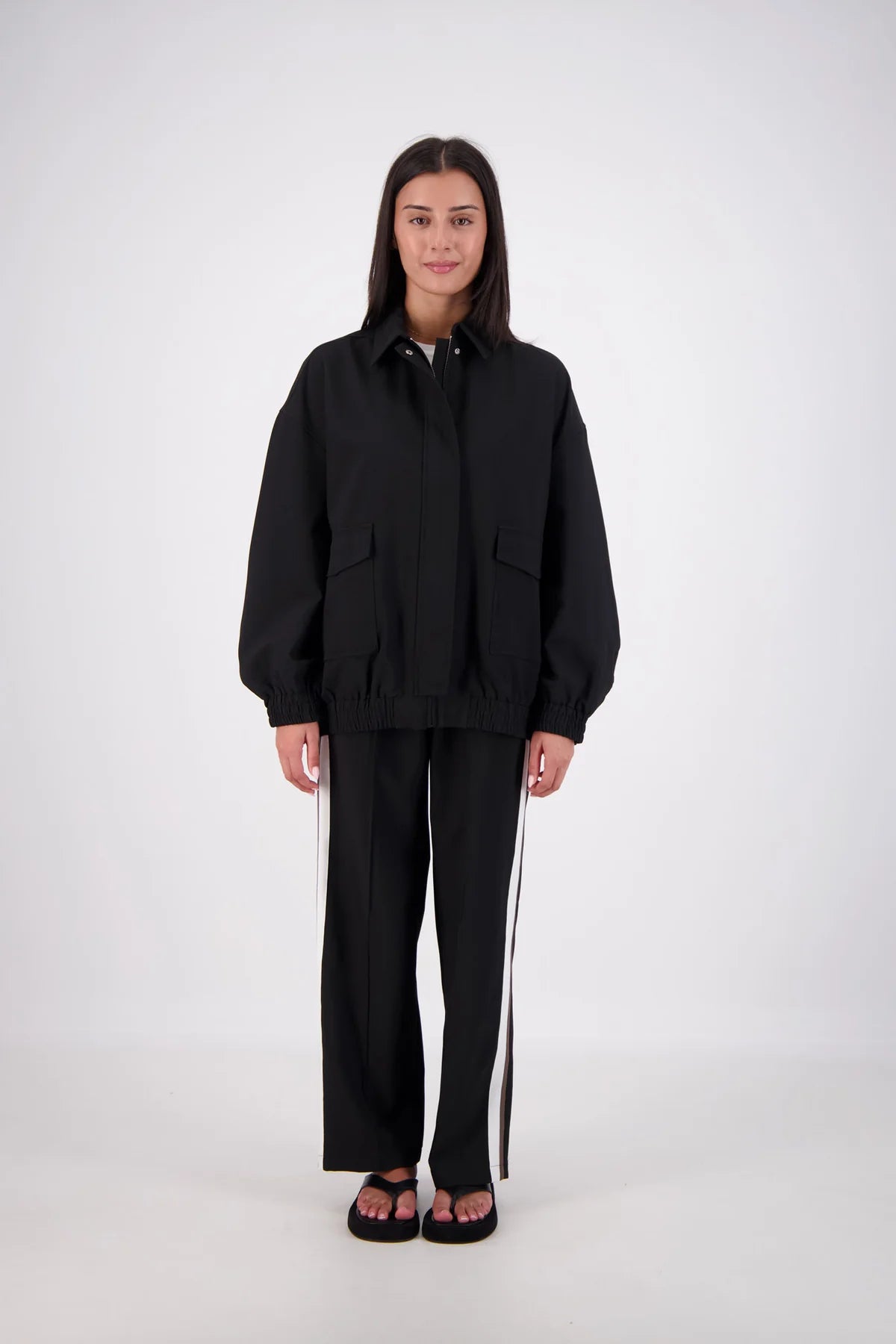 A woman stands against a light gray studio background wearing a black oversized zip-front jacket with front flap pockets and matching black wide-leg pants with white side stripes.