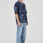 Person wearing a navy blue embroidered shirt and light blue jeans on a white background