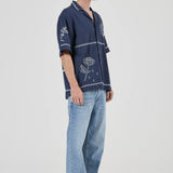 Person wearing a navy blue embroidered shirt and light blue jeans on a white background