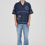 A model stands against a light gray studio background wearing a navy blue short-sleeve button-up shirt with white embroidered floral and circular patterns, paired with light blue jeans and gray sneakers.