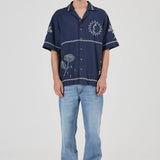 A model stands against a light gray studio background wearing a navy blue short-sleeve button-up shirt with white embroidered floral and circular patterns, paired with light blue jeans and gray sneakers.