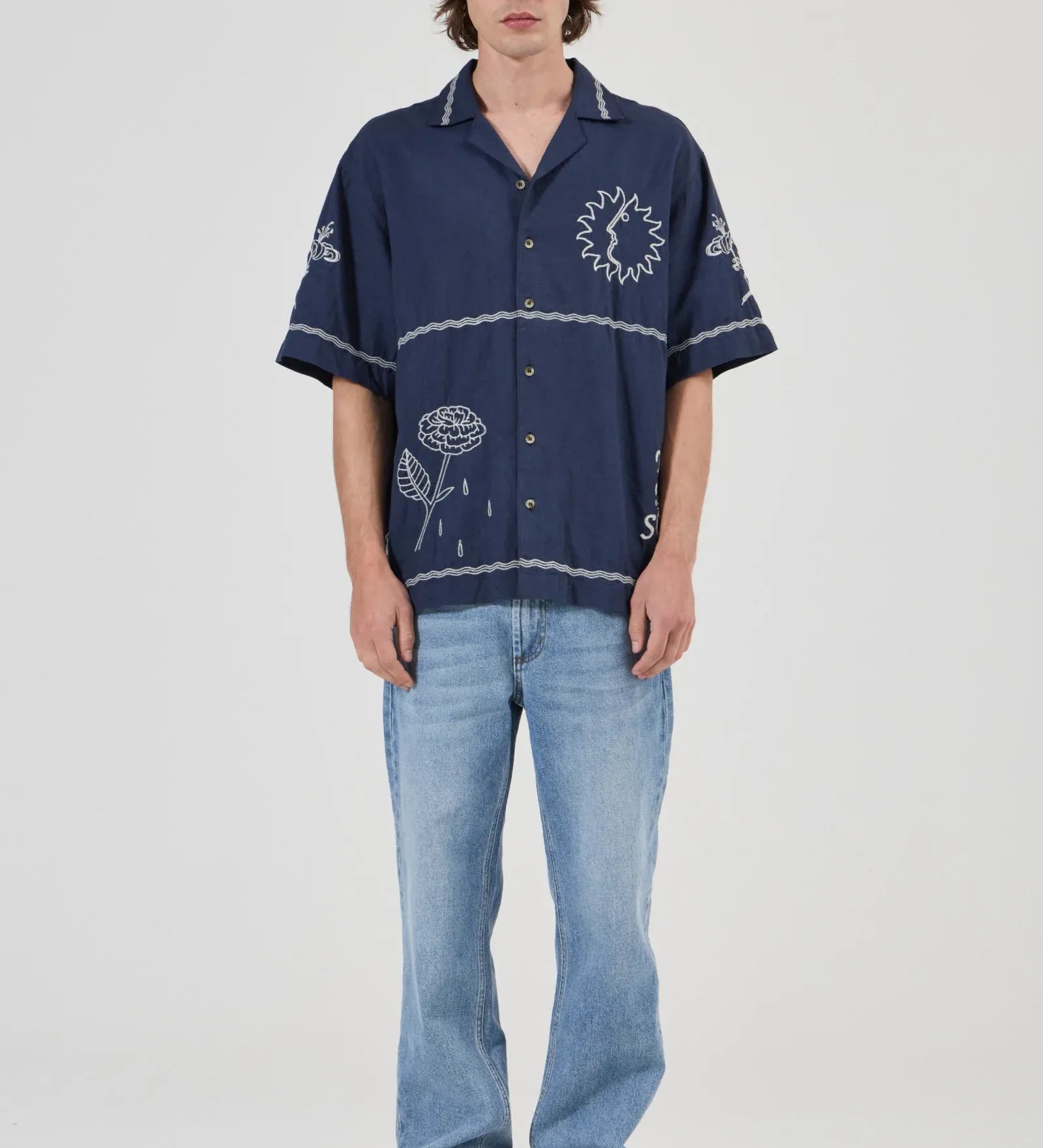 A model stands against a light gray studio background wearing a navy blue short-sleeve button-up shirt with white embroidered floral and circular patterns, paired with light blue jeans and gray sneakers.