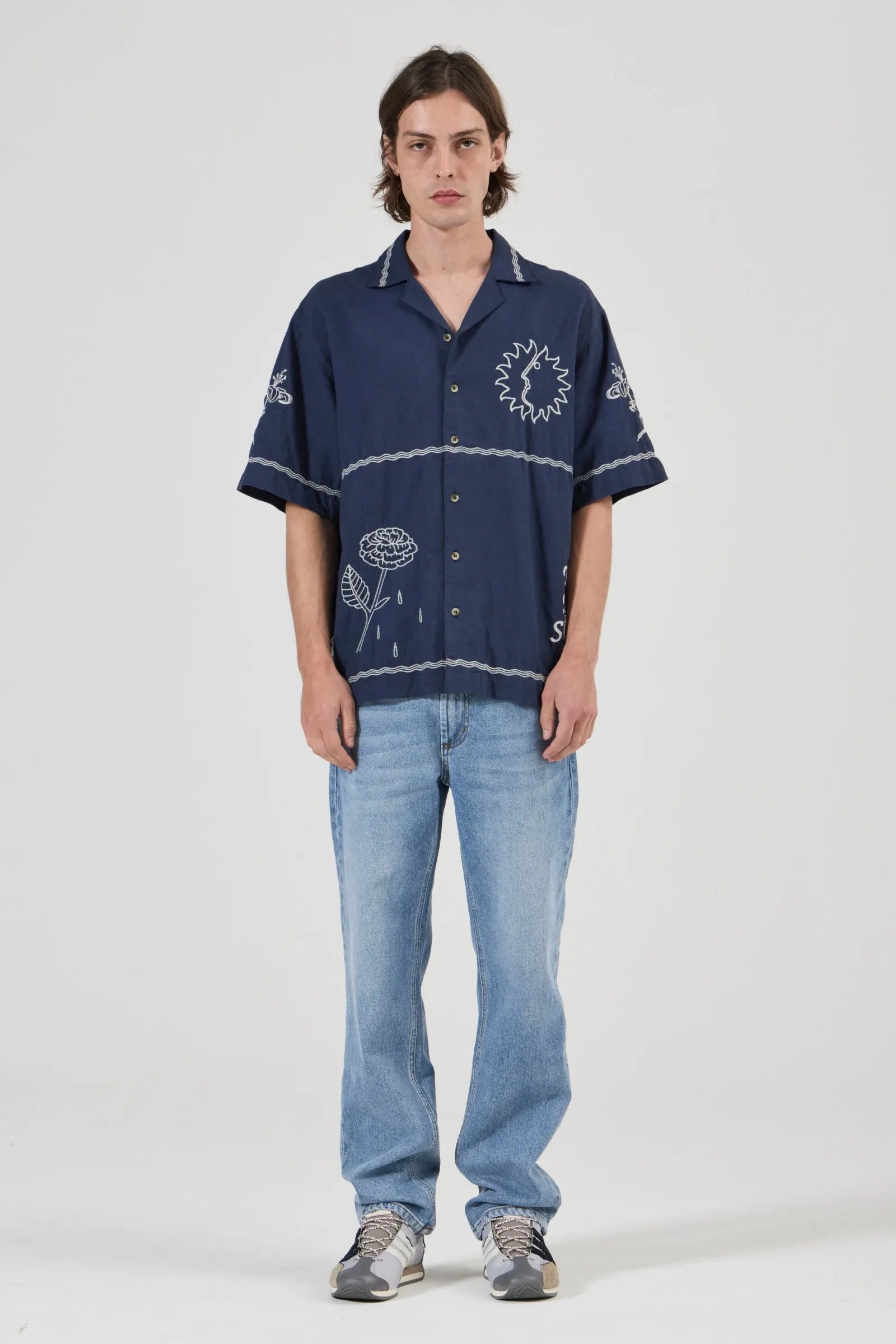 A model stands against a light gray studio background wearing a navy blue short-sleeve button-up shirt with white embroidered floral and circular patterns, paired with light blue jeans and gray sneakers.