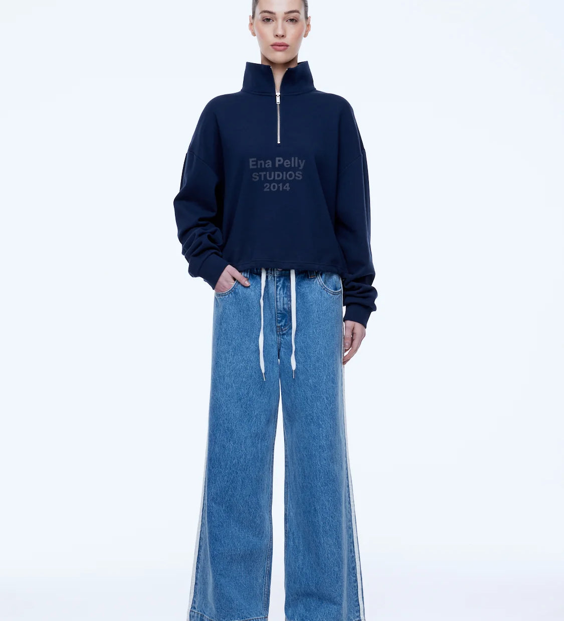 A model stands against a white studio background wearing a navy blue half-zip sweatshirt with white text reading 'ENA PELLY STUDIOS 2014' on the chest, paired with wide-leg blue jeans.