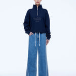 A model stands against a white studio background wearing a navy blue half-zip sweatshirt with white text reading 'ENA PELLY STUDIOS 2014' on the chest, paired with wide-leg blue jeans.