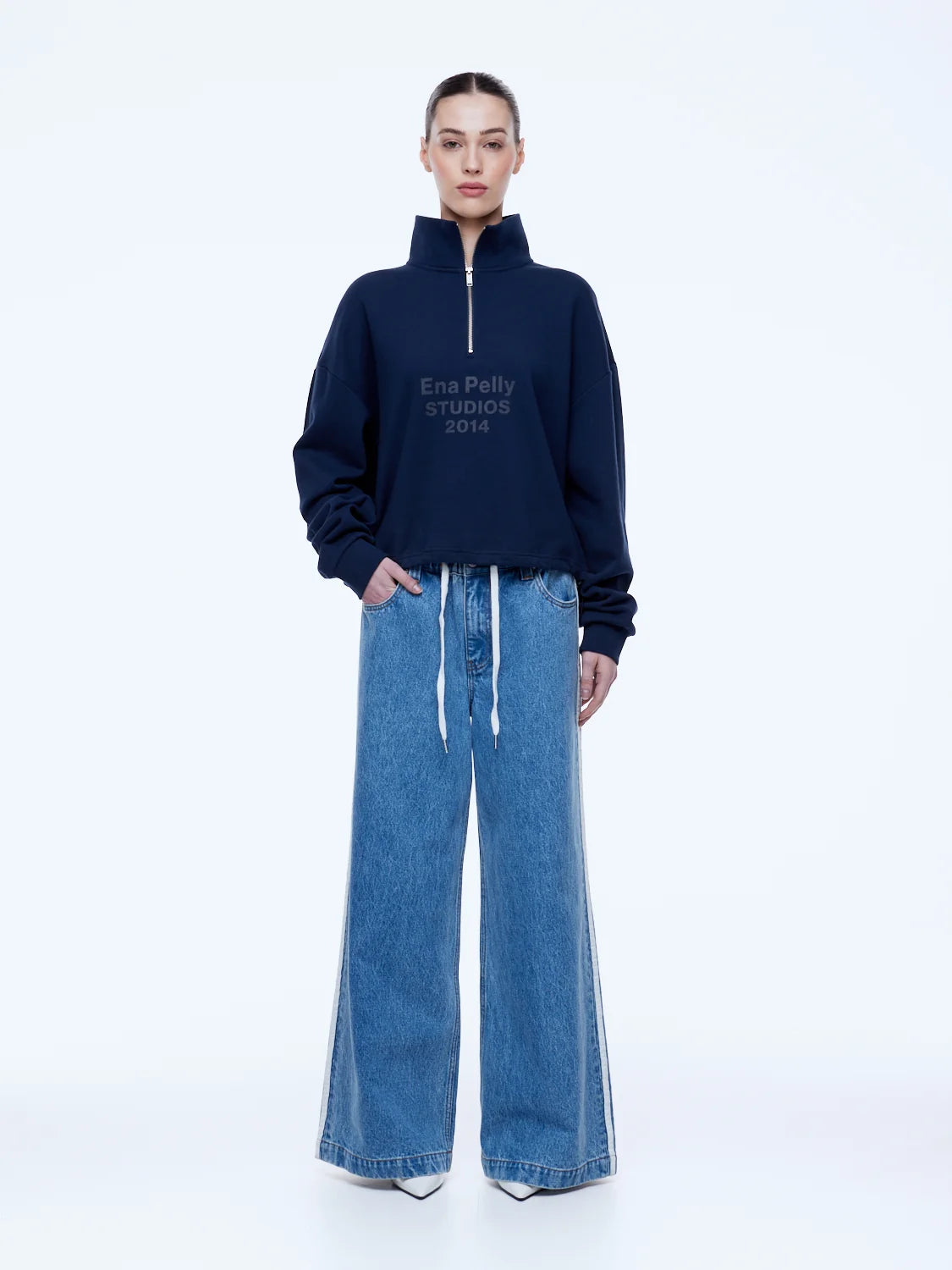 A model stands against a white studio background wearing a navy blue half-zip sweatshirt with white text reading 'ENA PELLY STUDIOS 2014' on the chest, paired with wide-leg blue jeans.
