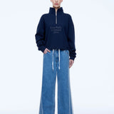 A model stands against a white studio background wearing a navy blue half-zip sweatshirt with white text reading 'ENA PELLY STUDIOS 2014' on the chest, paired with wide-leg blue jeans.
