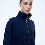 A woman wearing a navy blue quarter-zip sweatshirt with 'Ena Pelly STUDIOS 2014' printed in dark gray on the front, photographed against a clean white studio background.