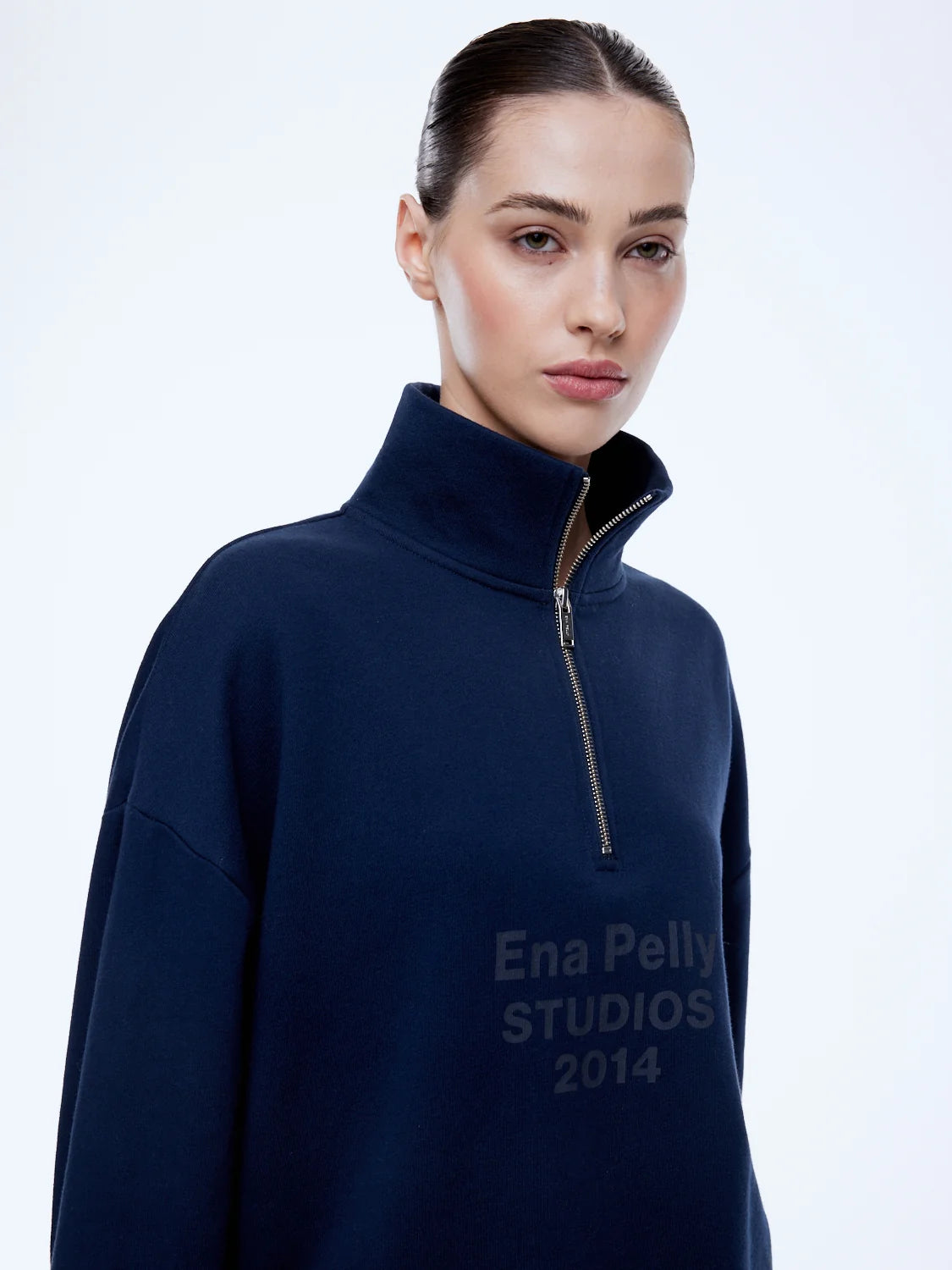 A woman wearing a navy blue quarter-zip sweatshirt with 'Ena Pelly STUDIOS 2014' printed in dark gray on the front, photographed against a clean white studio background.