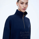 A woman wearing a navy blue quarter-zip sweatshirt with 'Ena Pelly STUDIOS 2014' printed in dark gray on the front, photographed against a clean white studio background.