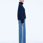 A model shown in profile wearing a dark navy blue mock-neck knit sweater and wide-leg blue denim jeans with white side stripes, against a plain white studio background.