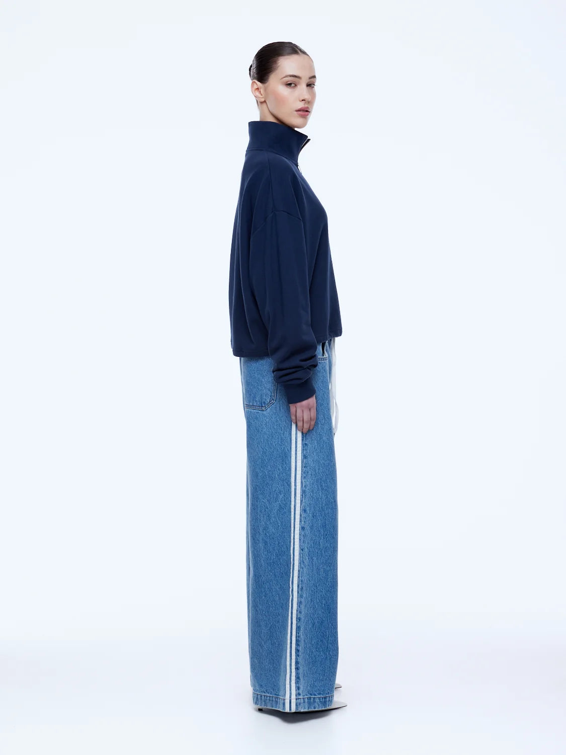 A model shown in profile wearing a dark navy blue mock-neck knit sweater and wide-leg blue denim jeans with white side stripes, against a plain white studio background.