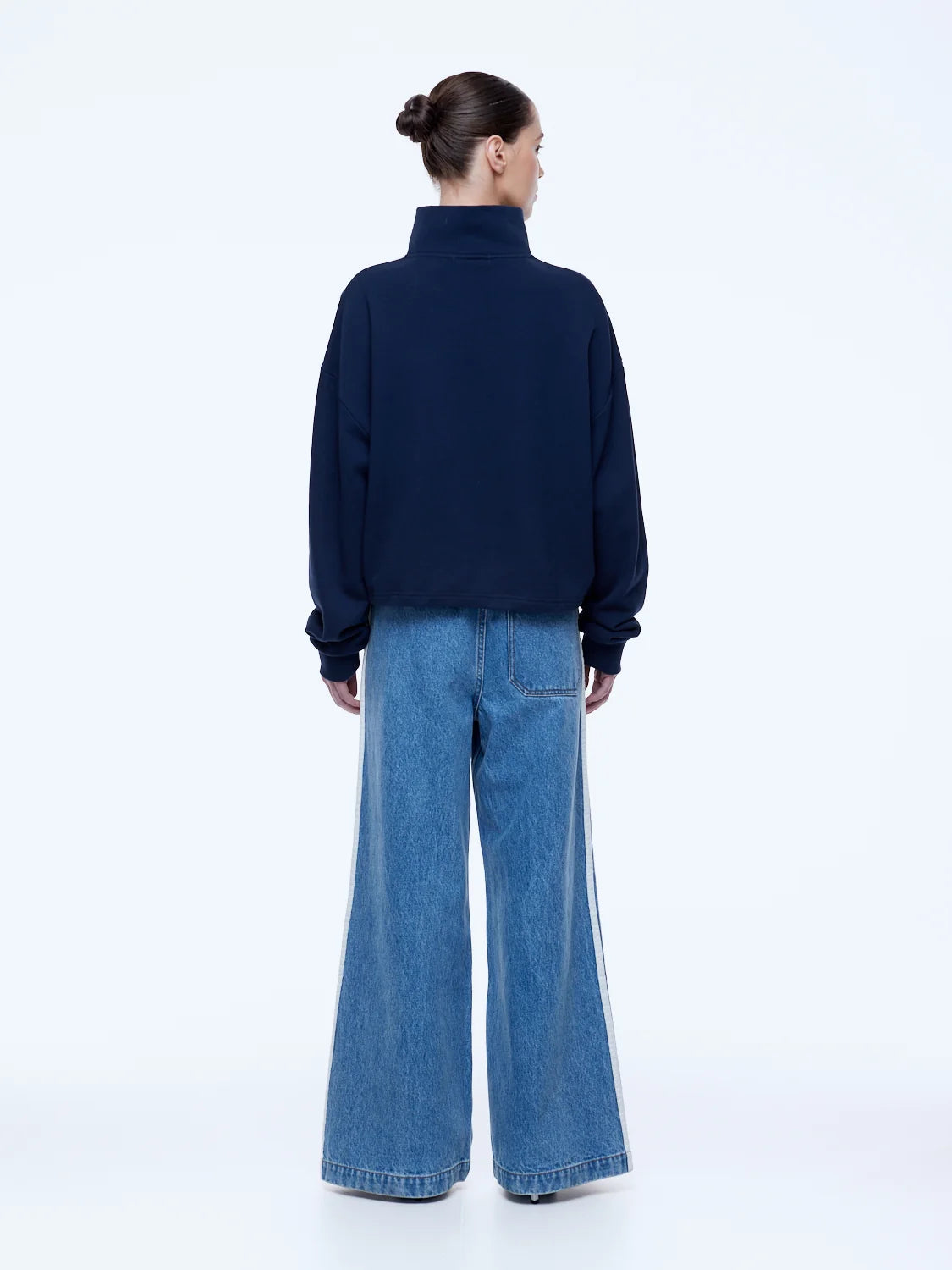 A model shown from the back wearing a dark navy blue mock-neck sweatshirt and wide-leg blue denim jeans with white side stripes, against a plain white studio background.