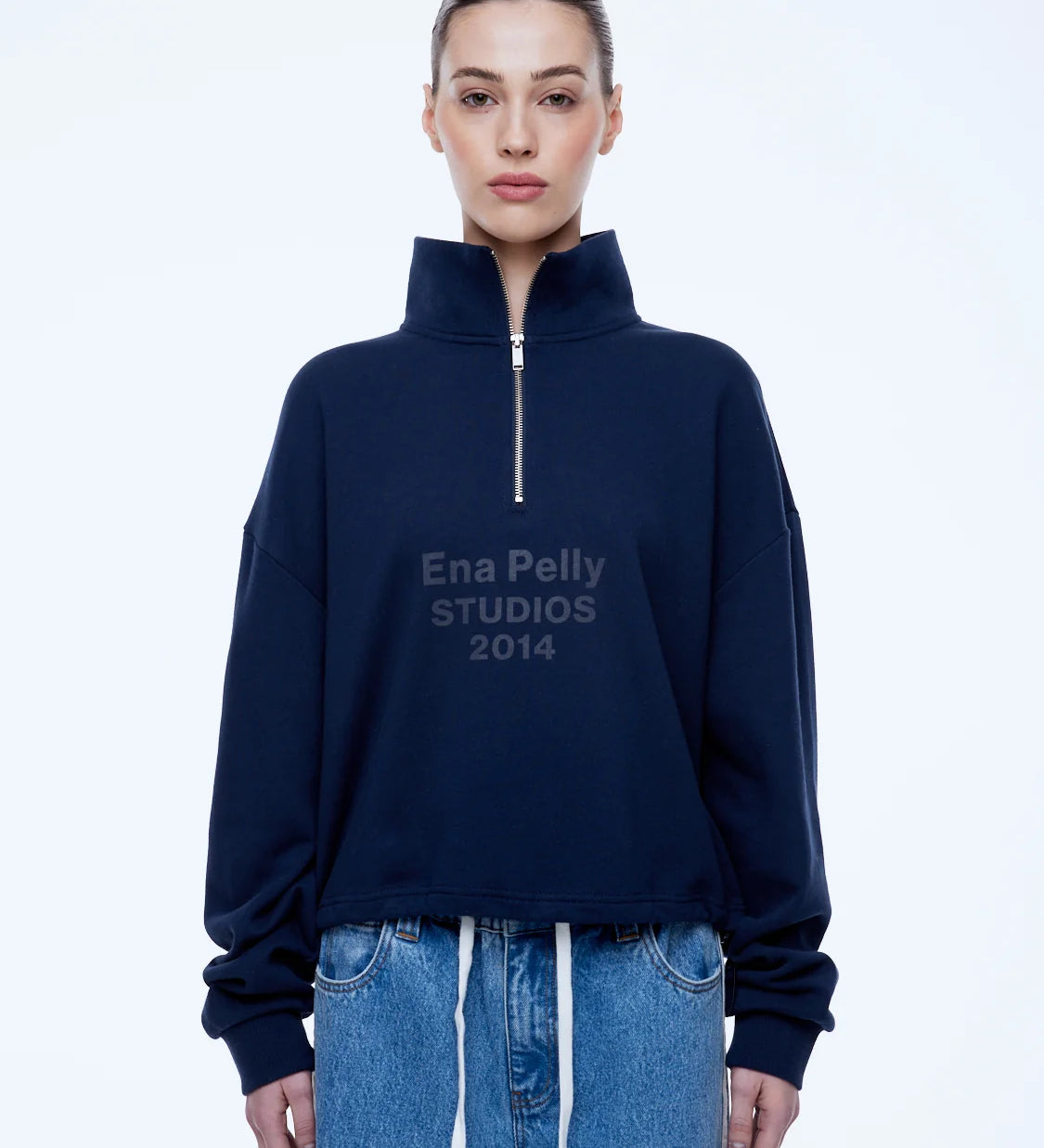 A model wearing a navy blue half-zip sweatshirt with 'Ena Pelly STUDIOS 2014' printed on the chest, paired with light blue denim jeans, against a plain white studio background.