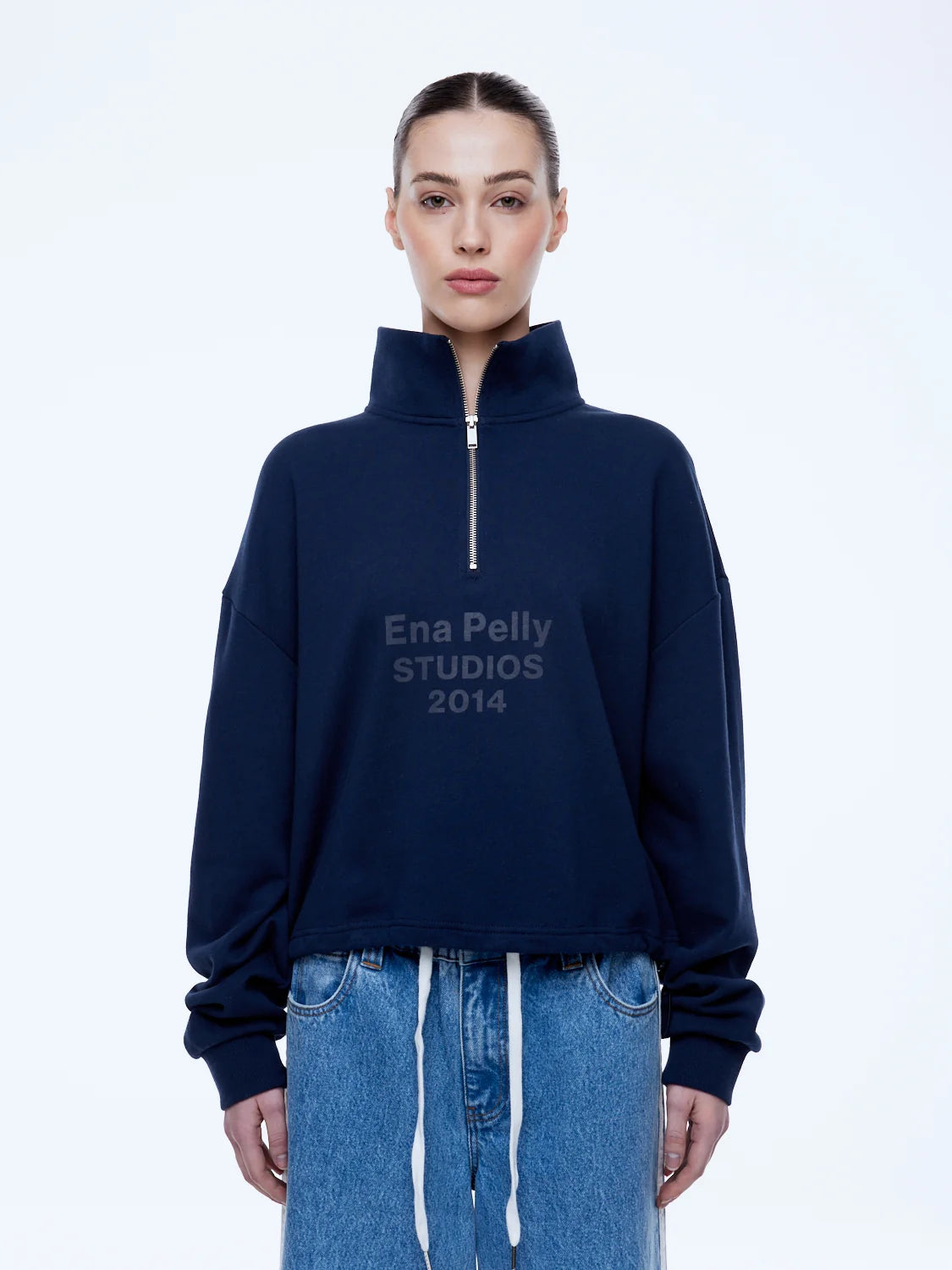 A model wearing a navy blue half-zip sweatshirt with 'Ena Pelly STUDIOS 2014' printed on the chest, paired with light blue denim jeans, against a plain white studio background.