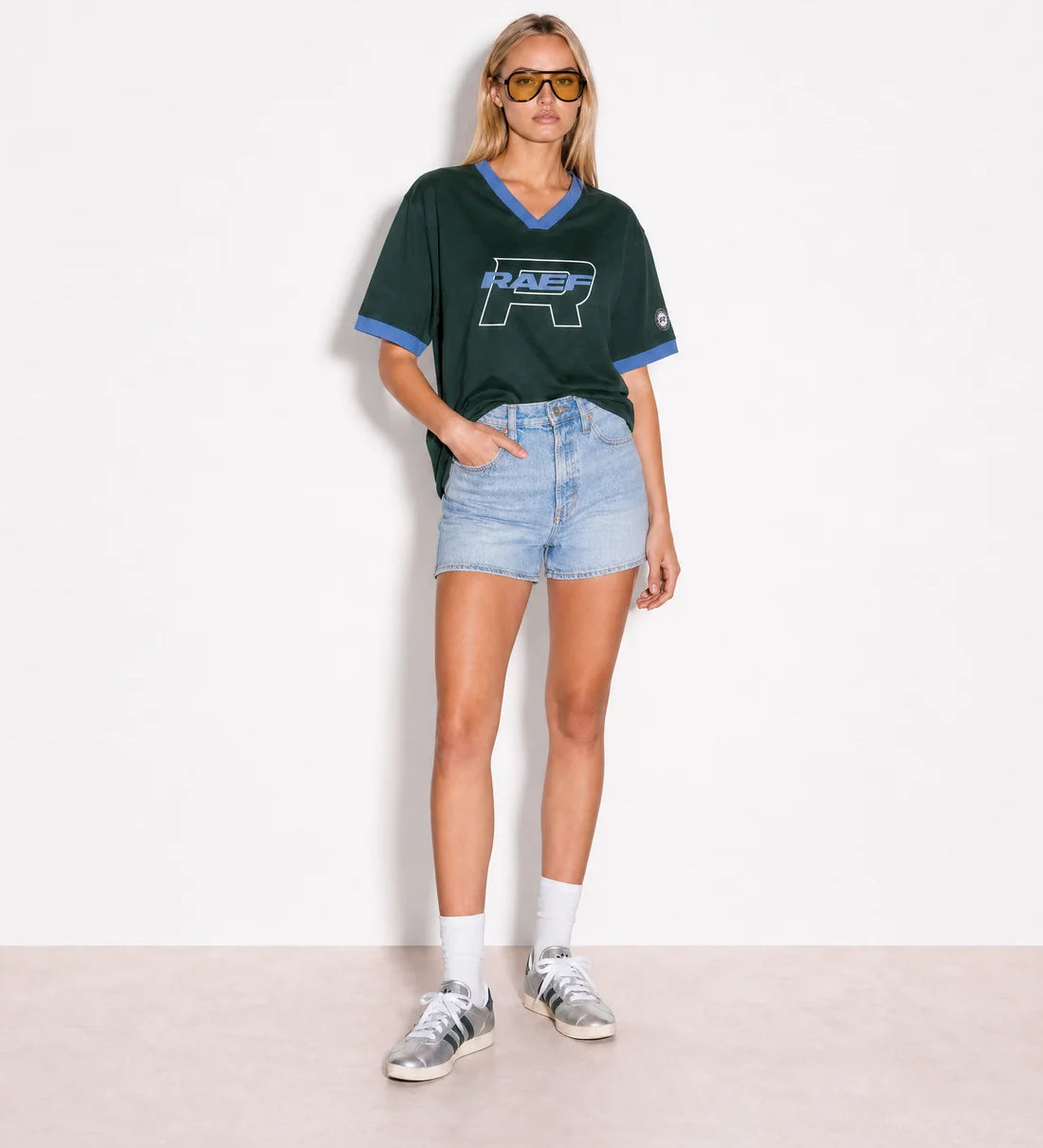 A model stands against a plain white wall wearing a dark green short-sleeve V-neck t-shirt with light blue trim and a large 'R.A.