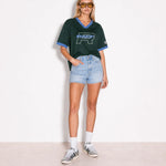 A model stands against a plain white wall wearing a dark green short-sleeve V-neck t-shirt with light blue trim and a large 'R.A.