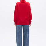 A model viewed from the back wearing a bright red mock-neck knit sweater and wide-leg blue jeans against a plain white studio background.