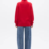 A model viewed from the back wearing a bright red mock-neck knit sweater and wide-leg blue jeans against a plain white studio background.