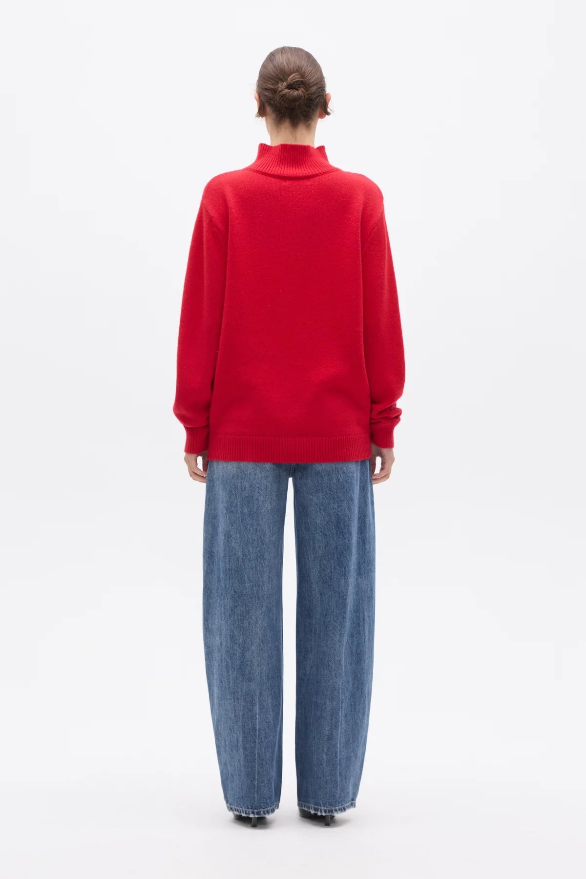 A model viewed from the back wearing a bright red mock-neck knit sweater and wide-leg blue jeans against a plain white studio background.