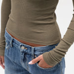 A close-up of a person wearing a fitted ribbed long-sleeve top in taupe/olive-brown and blue jeans, with one hand in a pocket, against a plain light background.