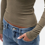 A close-up of a person wearing a fitted ribbed long-sleeve top in taupe/olive-brown and blue jeans, with one hand in a pocket, against a plain light background.