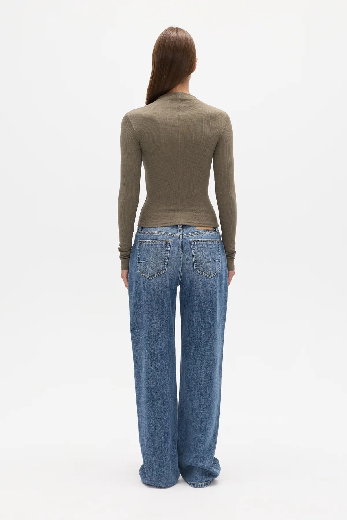 A woman viewed from the back wearing a fitted olive-brown long-sleeve mock-neck top and wide-leg blue jeans against a plain white studio background.