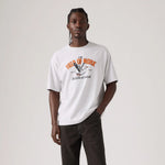 A man modeling a white graphic t-shirt with an orange and black bird design on the chest, paired with dark brown pants, against a plain light gray background.