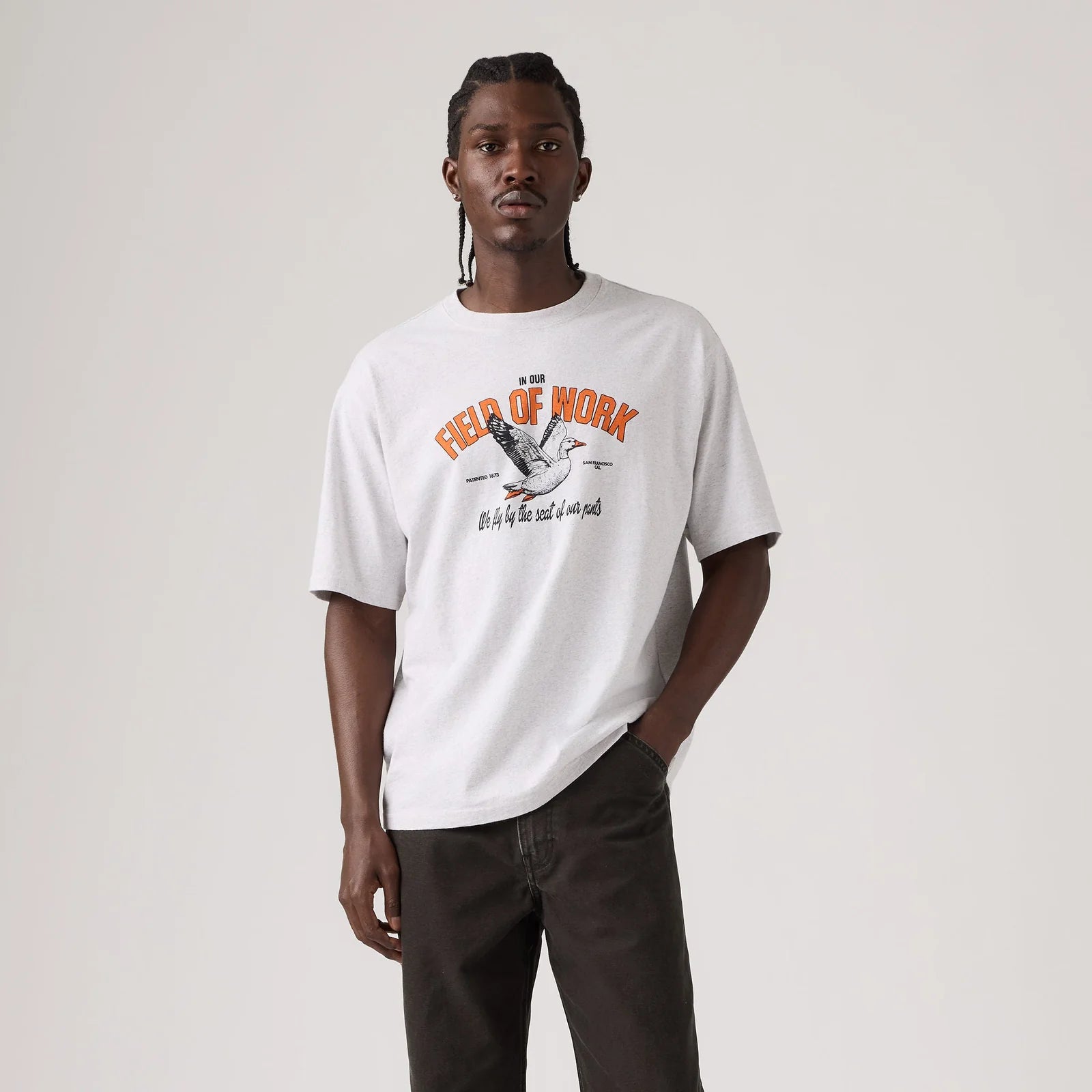 A man modeling a white graphic t-shirt with an orange and black bird design on the chest, paired with dark brown pants, against a plain light gray background.