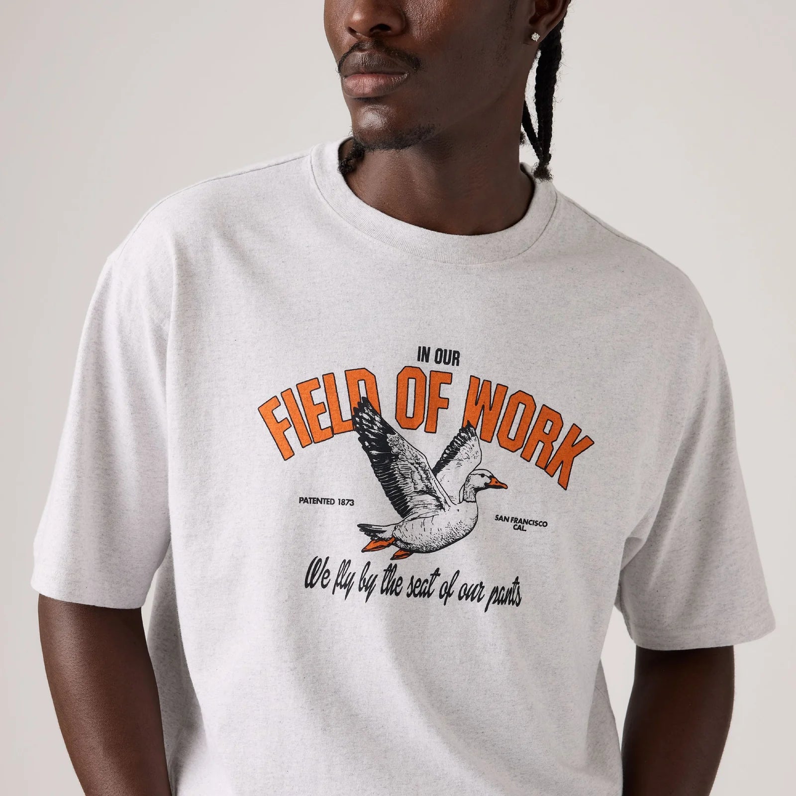 A man wearing a light gray crewneck t-shirt with a vintage-style graphic of a goose and the text 'IN OUR FIELD OF WORK We fly by the seat of our pants'.