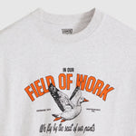 A close-up of a light heather gray Levi's t-shirt featuring a vintage-style graphic print with orange text reading 'FIELD OF WORK', a flying duck, and black script text below.