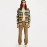 A model stands against a plain white studio background wearing a striped knit cardigan with yellow, green, and blue tones over a cream shirt, paired with tan pants and white sneakers.