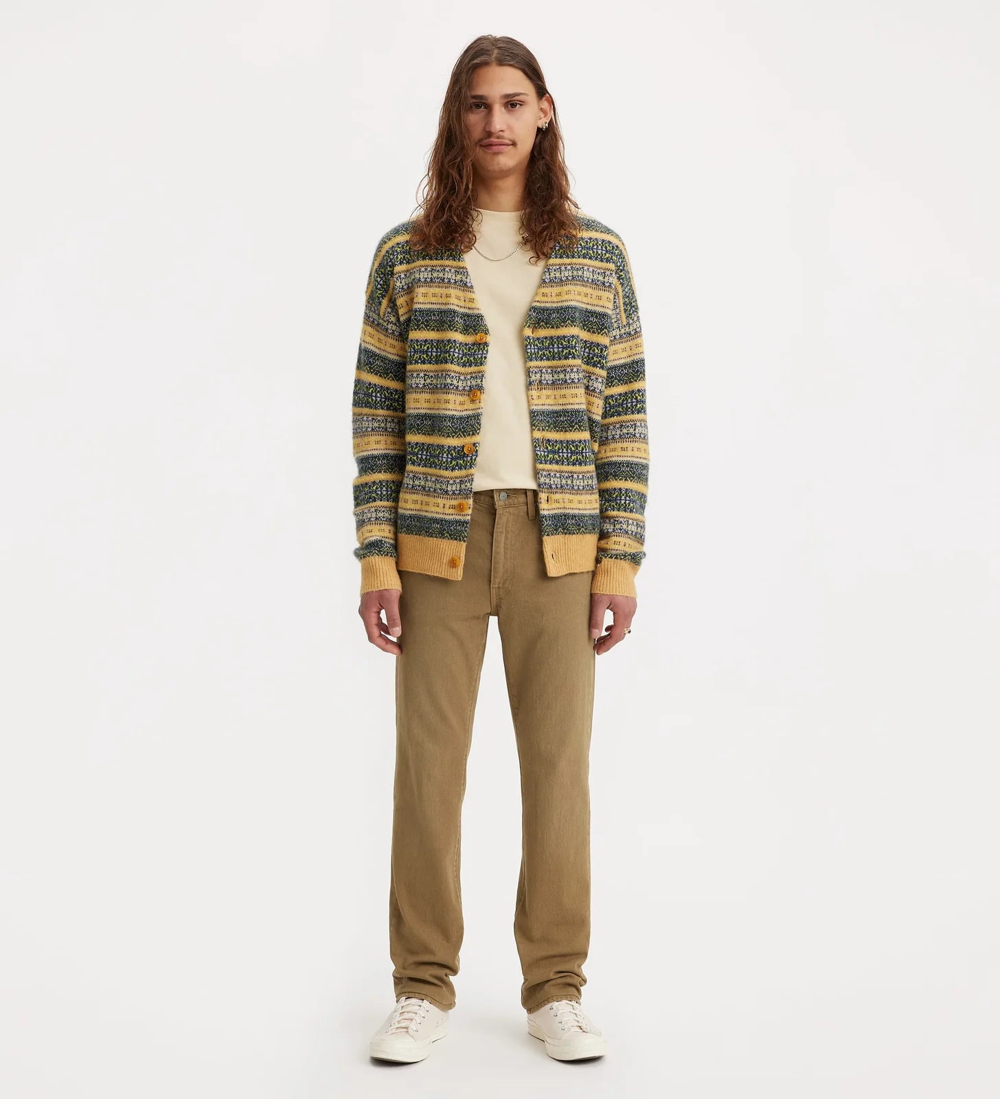 A model stands against a plain white studio background wearing a striped knit cardigan with yellow, green, and blue tones over a cream shirt, paired with tan pants and white sneakers.