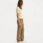 A man with long brown hair is shown in profile wearing tan straight-leg jeans, a cream t-shirt, and white sneakers against a plain white studio background.