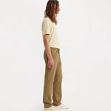 A man with long brown hair is shown in profile wearing tan straight-leg jeans, a cream t-shirt, and white sneakers against a plain white studio background.
