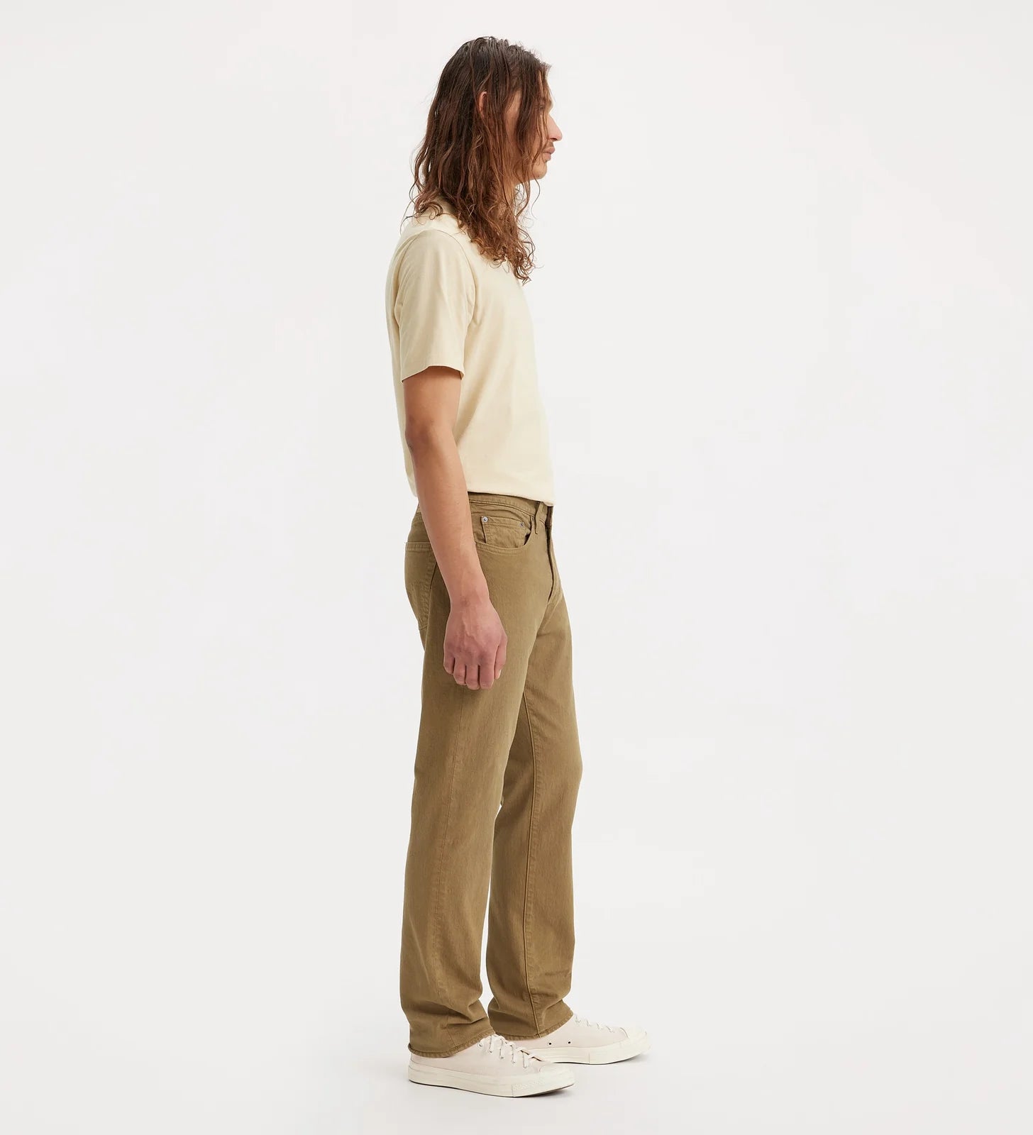 A man with long brown hair is shown in profile wearing tan straight-leg jeans, a cream t-shirt, and white sneakers against a plain white studio background.