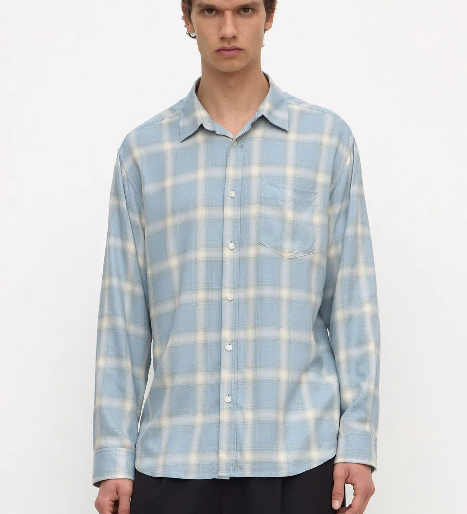 A man wearing a light blue and white plaid long-sleeve button-up shirt with dark trousers, standing against a plain white studio background.