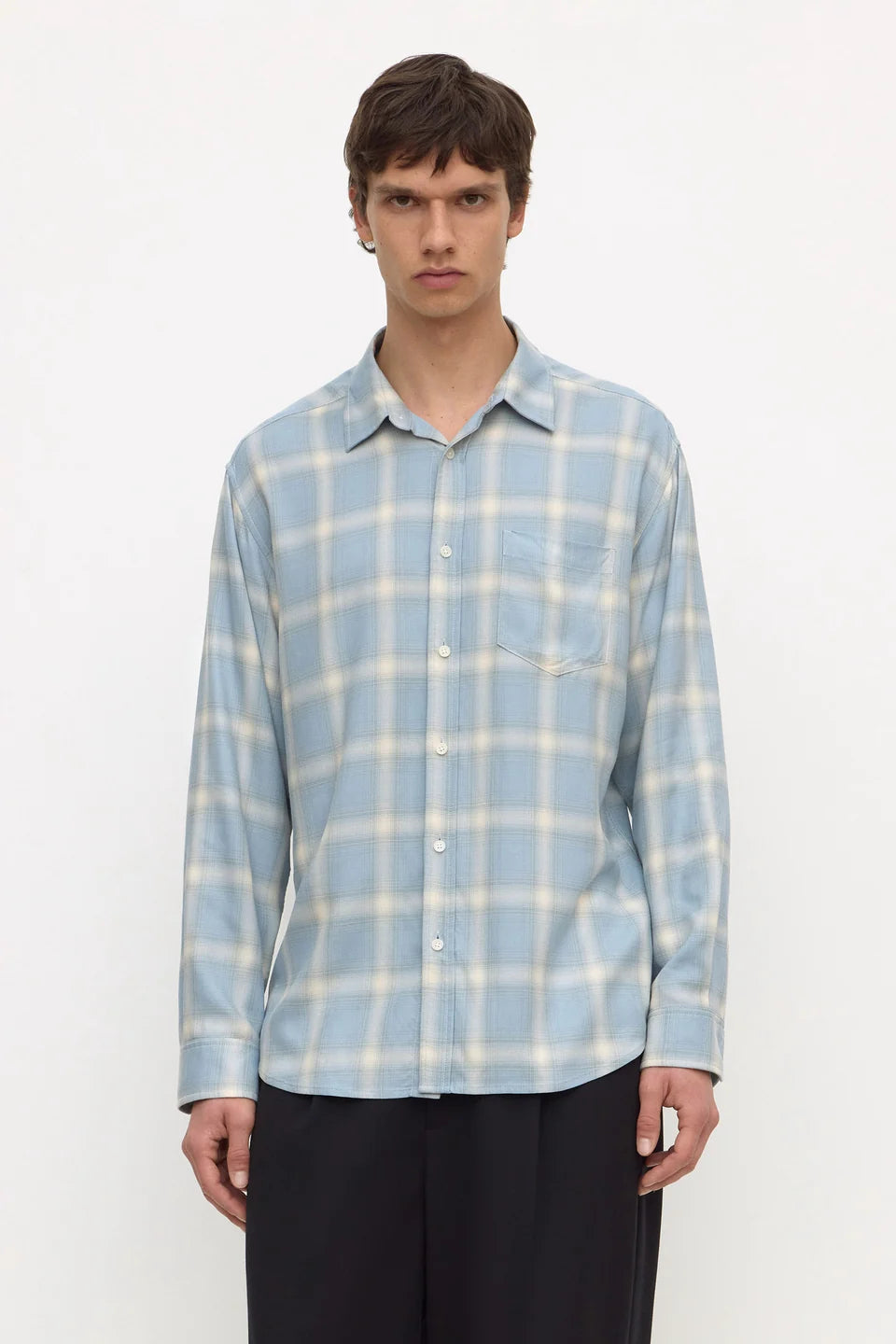 A man wearing a light blue and white plaid long-sleeve button-up shirt with dark trousers, standing against a plain white studio background.