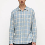 A man wearing a light blue and white plaid long-sleeve button-up shirt with dark trousers, standing against a plain white studio background.