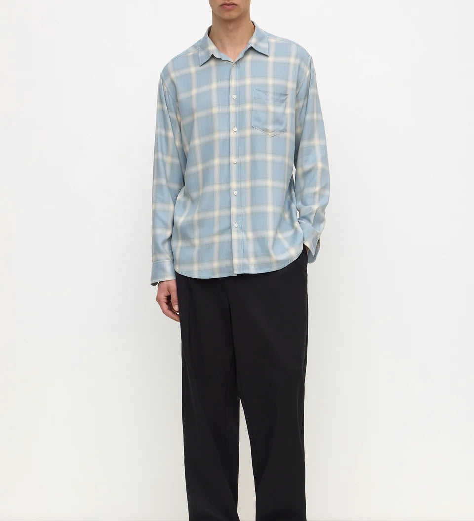 A man modeling a light blue and white plaid long-sleeve button-up shirt with black trousers, standing against a plain white studio background.