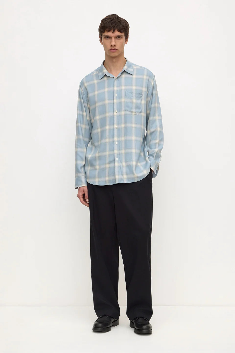 A man modeling a light blue and white plaid long-sleeve button-up shirt with black trousers, standing against a plain white studio background.
